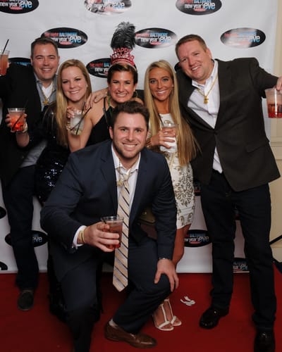 Six friends in party beads and a New Year's tiara raise their drinks on the red carpet in front of a New Year's Eve backdrop.