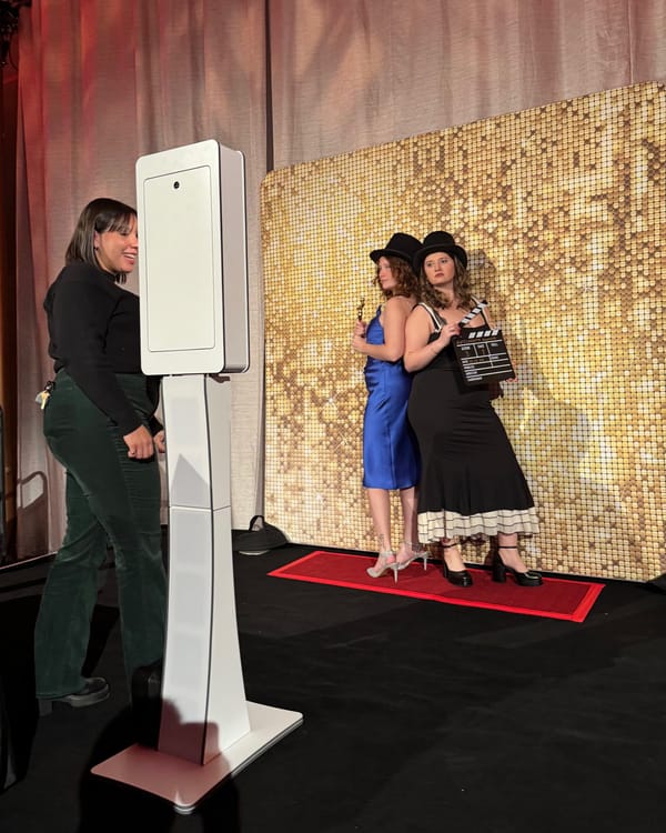 Two guests in top hats pose with a clapperboard on a red carpet in front of a gold sequin wall, next to a white photo booth kiosk.