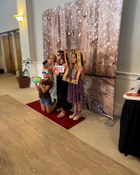 Four girls in party sunglasses hold up props and a frame on the red carpet in front of a rustic wood backdrop.