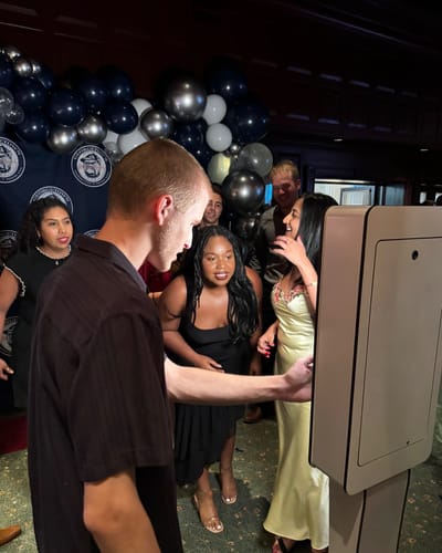 Guests laugh as one taps the open-air booth's screen at a university gala, with a balloon arch and step-and-repeat behind them.