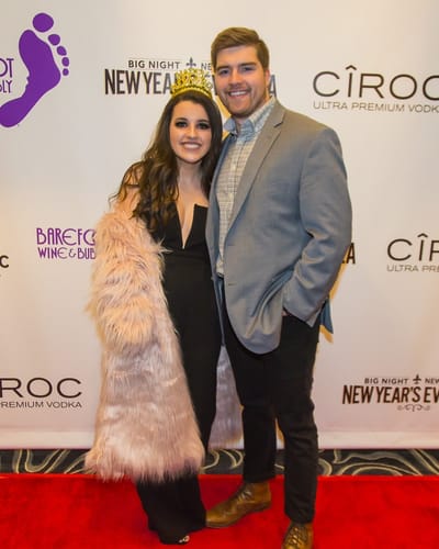 A couple, she in a party crown and faux-fur coat, smile on the red carpet at a New Year's Eve gala sponsor wall.