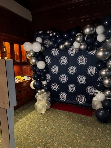 A navy, silver and white balloon arch framing a university step-and-repeat backdrop and red carpet.