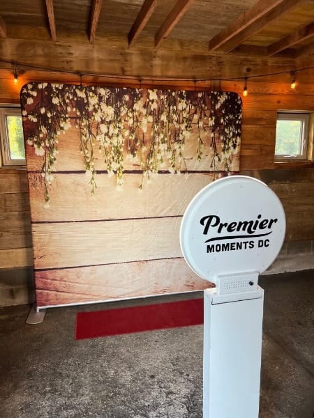 A wood backdrop hung with white flowers, a red carpet and a Premier Moments DC kiosk in a barn.