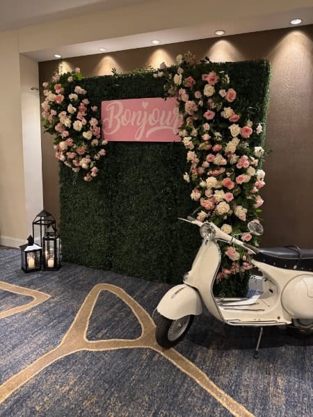 A greenery wall with pink and white roses and a Bonjour sign, with lanterns and a vintage scooter.