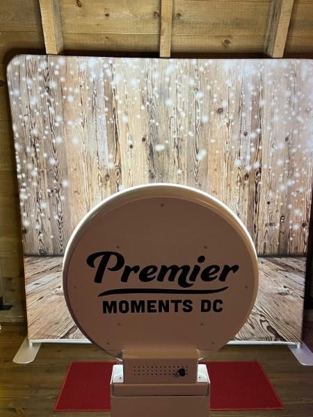 A rustic wood backdrop with sparkling lights behind a Premier Moments DC kiosk and red carpet.